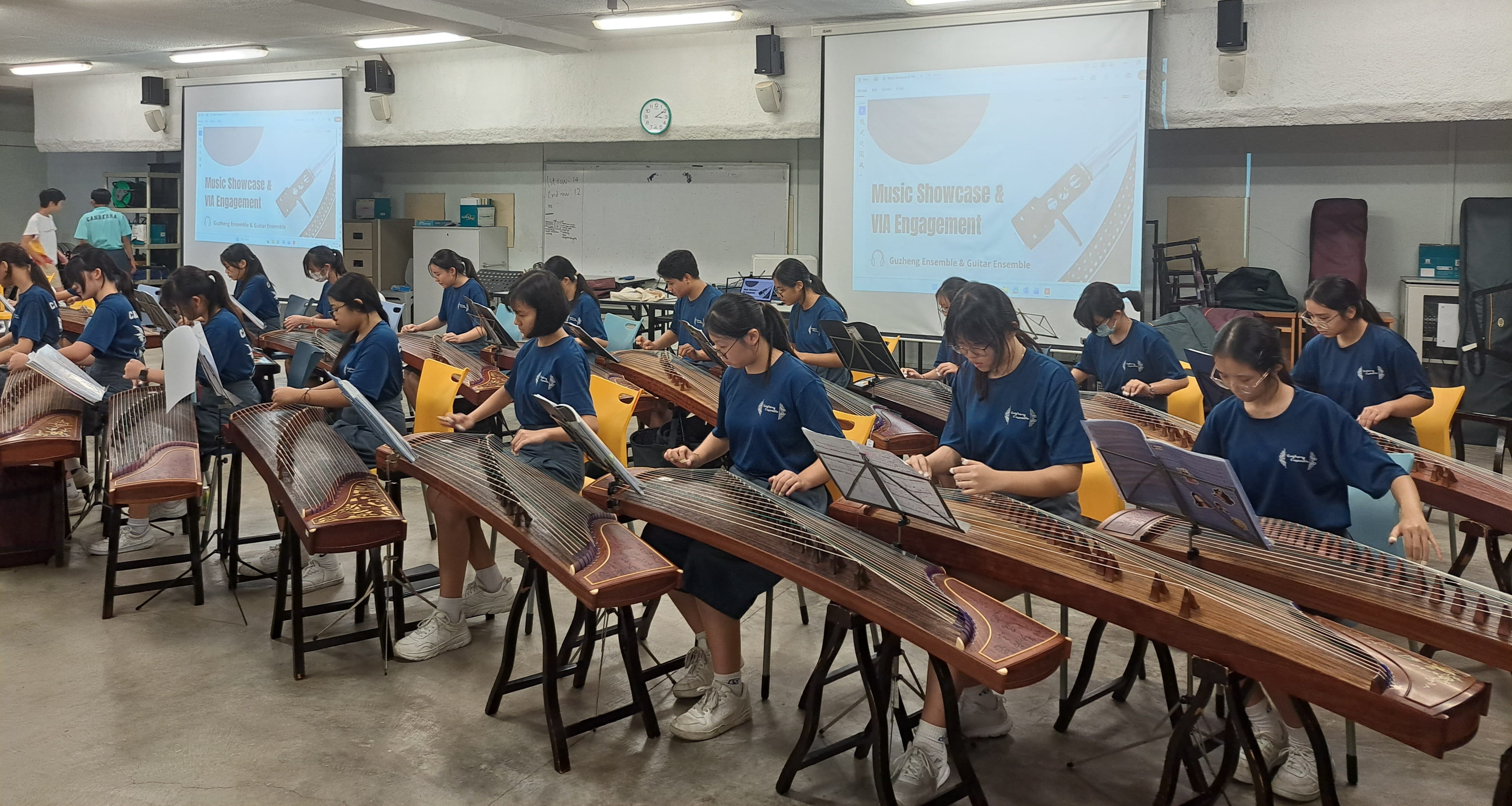 A group of approximately 20 students dressed in matching navy blue T-shirts playing the guzheng (Chinese zither) during a rehearsal or practice session. The students are seated in rows behind their instruments, reading from music scores on stands. In the background, two projection screens display a presentation slide titled 'Music Showcase & VIA Engagement' by the Guzheng Ensemble & Guitar Ensemble. The setting appears to be a school music room or hall.